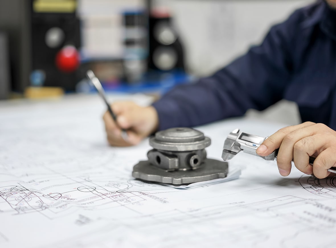 A person uses a caliper to measure a metal part and holds a pen over engineering blueprints, indicating an industrial design process.