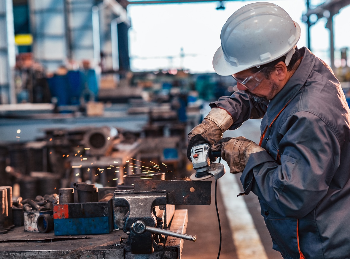 A worker in safety gear operates an angle grinder on metal in an industrial workshop, sparks flying over a cluttered workbench.