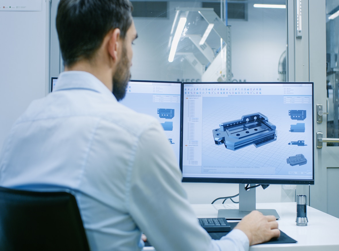 A man in a white shirt works at a desk with dual monitors showing 3D CAD models of a mechanical part in a modern office or lab.