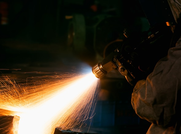 A person wearing protective clothing uses a torch or welding tool, sending bright orange sparks flying in an industrial workshop.
