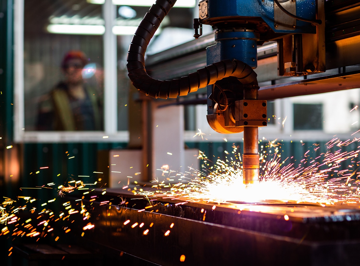 A CNC machine slices metal, sending off bright sparks in an industrial workshop; a person in safety gear stands behind a glass window.